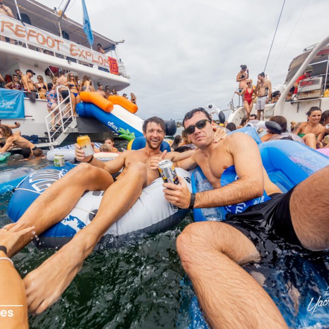 A group of people on and around a boat are enjoying a party on the water. Two men smiling and holding drinks are floating in an inflatable pool toy. Other partygoers are visible in the background on the boat and in the water. Banners on the boat read “The Yacht Social Club Event Boat Charters.”