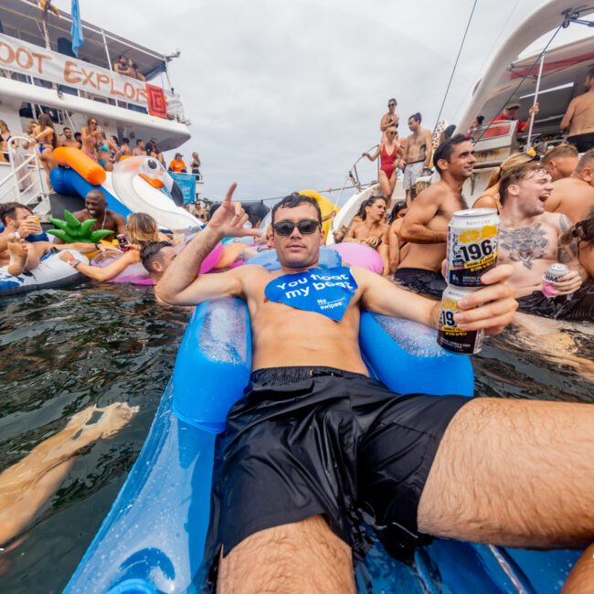 A man in sunglasses and a blue life vest floats on an inflatable raft, holding a drink can and making a peace sign. Around him, several people are on inflatables and swimming near large party boats, enjoying The Yacht Social Club's lively social gathering.