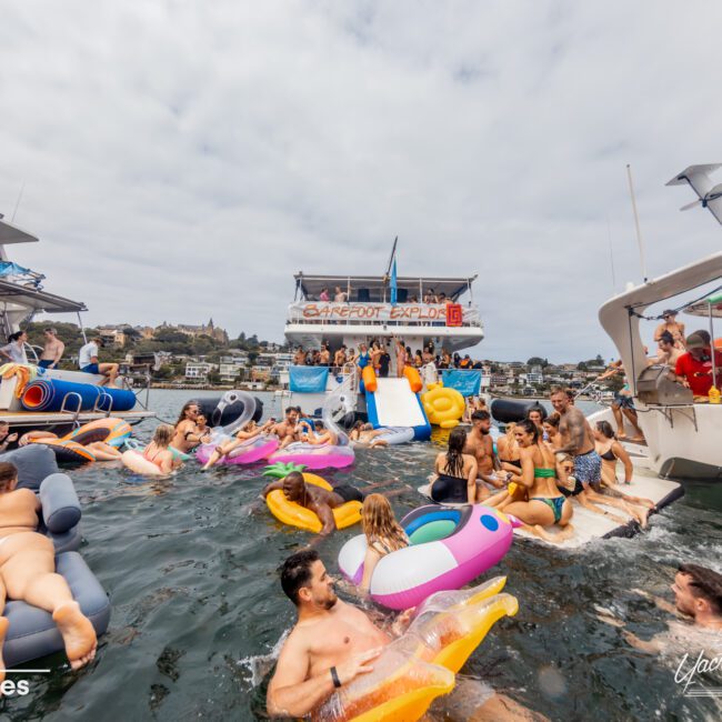 A lively boat party hosted by The Yacht Social Club is taking place with numerous people enjoying themselves in and around the water. Some are lounging on colorful inflatables, while others are swimming. Several boats are docked nearby, adorned with festive decorations and banners.