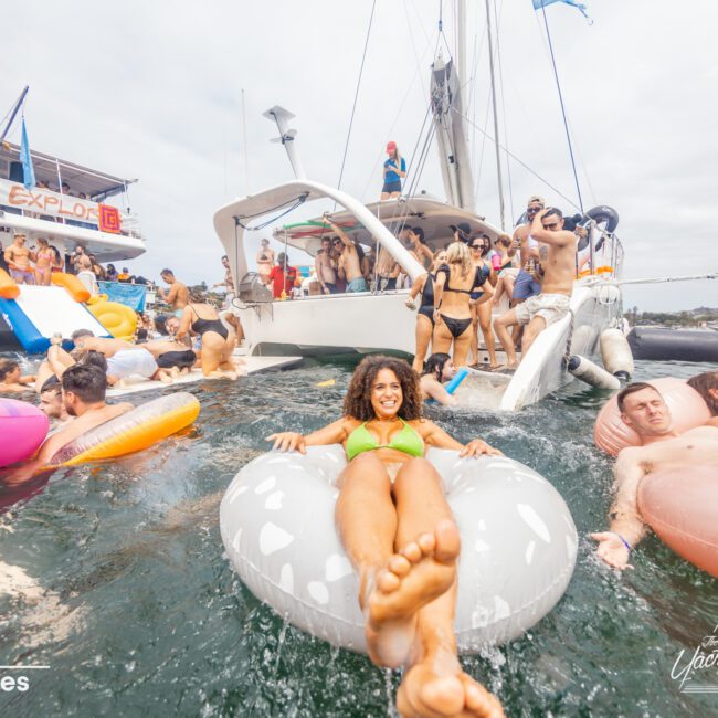 A lively scene of a boat party features people in swimsuits enjoying the water on various pool floats. A woman smiles while relaxing on a white float in the foreground. The background shows a boat with more partygoers and inflatable floats, exemplifying the fun at The Yacht Social Club Sydney Boat Hire.