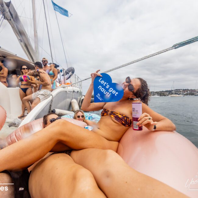 A group of people is enjoying a sunny day on a boat from The Yacht Social Club Sydney Boat Hire. A woman in the foreground reclines on an inflatable float, holding a drink and a "Let's get nauti" sign. The scene is lively with others lounging and socializing, water, and a cloudy sky in the background.