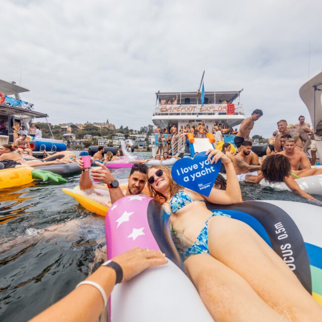 A lively boat party scene with people on inflatables in the water. A couple in the center holds signs, one reading "I love you a yacht." In the background, The Yacht Social Club's luxurious yacht stands out while people interact. The sky is cloudy, and the atmosphere is festive and joyful.