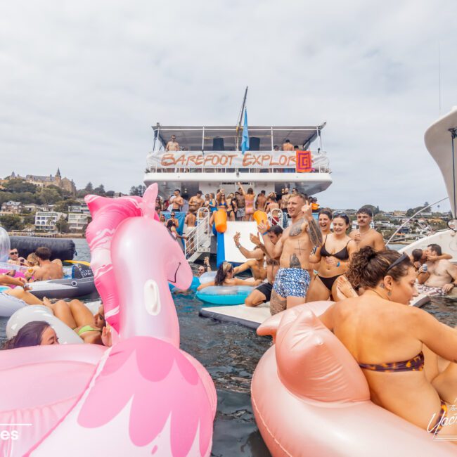 A group of people enjoying a boat party on the water, surrounded by various inflatable floats including a large pink flamingo. Two boats in the background, one with "BAREFOOT EXPLOR" written on it. People are socializing and swimming, showcasing the vibrant atmosphere of Boat Parties Sydney The Yacht Social Club.