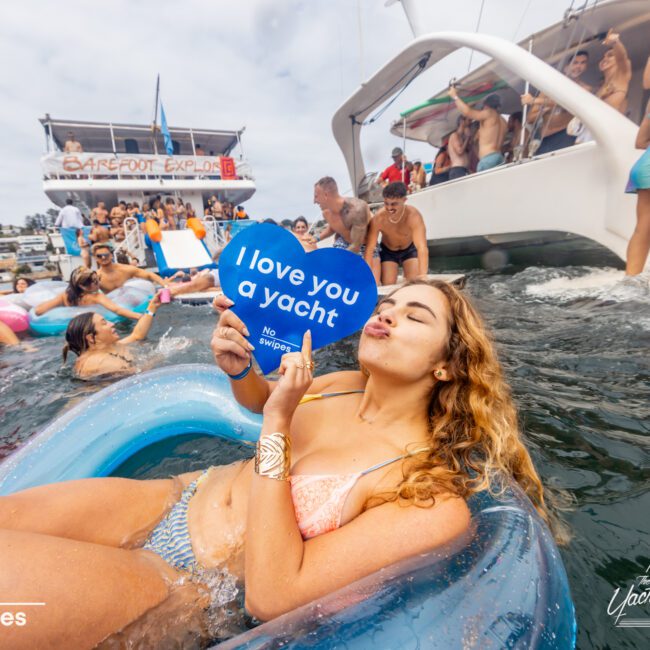 A woman in a bikini floats on an inflatable in the water, holding a sign that reads "I love you a yacht." Several people are partying on a luxury yacht rental in Sydney Harbour. The scene is lively with laughter, splashes, and colorful floats, capturing the vibrant spirit of The Yacht Social Club.