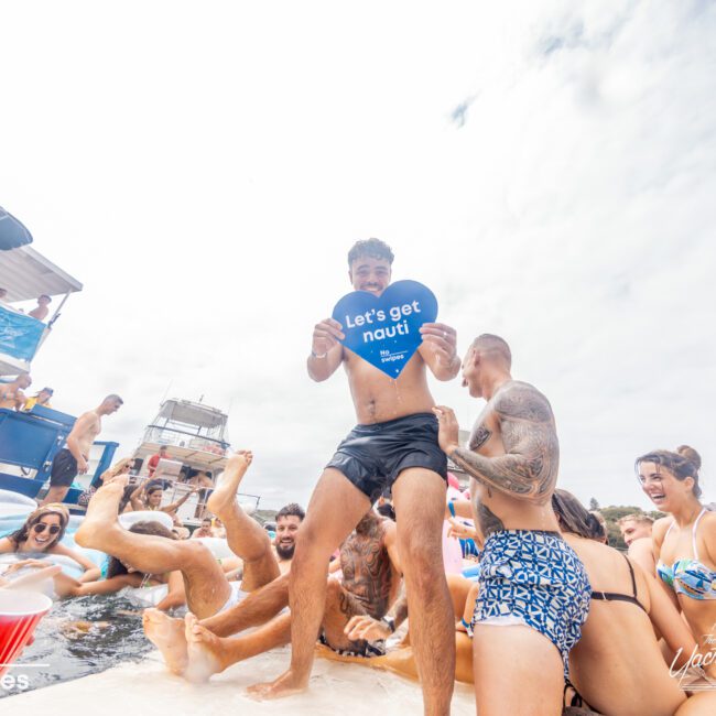 A crowd enjoys a boat party, with some standing on a float. One man in the center holds a heart-shaped sign that reads "Let's get nauti." Other boats are visible in the background, and people appear to be in swimsuits, having fun in the sun at The Yacht Social Club Event Boat Charters.