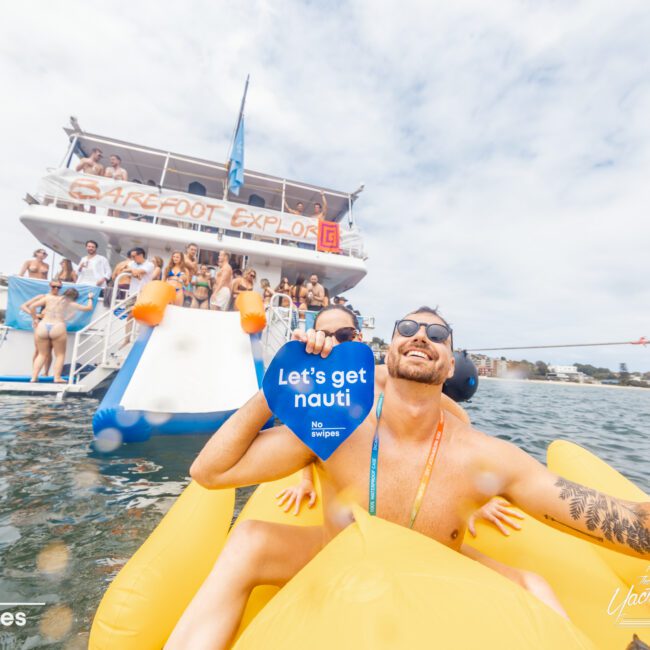 A lively boat party with people in swimsuits. In the foreground, a man in sunglasses holds a sign that reads "Let's get nauti." A large boat from The Yacht Social Club Event Boat Charters is in the background, with some people lounging on the deck and others enjoying Sydney Harbour.