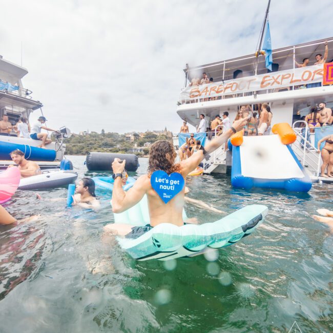 People are enjoying a party on boats and in the water. There is a woman in the foreground, wearing a blue shirt that reads "Let's get naval." She is riding a floating device and holding hands with someone. Other partygoers are seen around a large yacht from Luxury Yacht Rentals Sydney in the background.