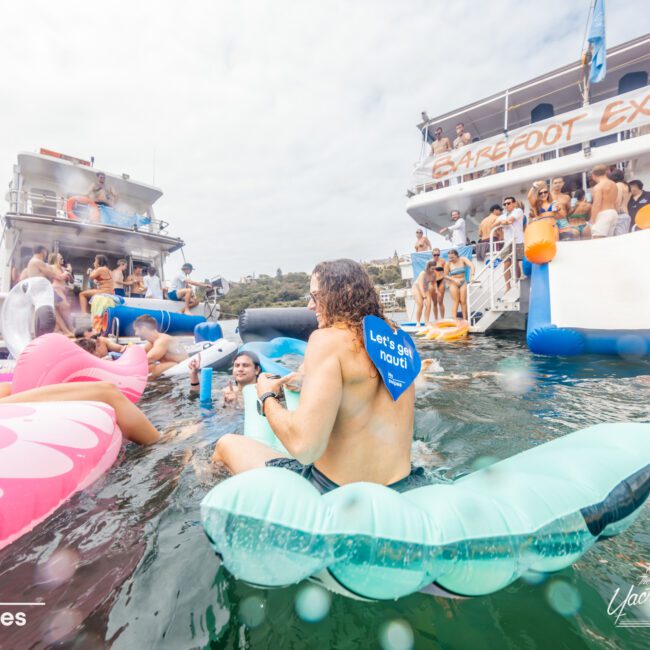 A crowded, lively scene with people enjoying a pool party on a large boat rented from "Luxury Yacht Rentals Sydney." Attendees are on various colorful inflatables in the water, while others are on the boat's deck and a slide leads into the water. The atmosphere appears festive and vibrant.