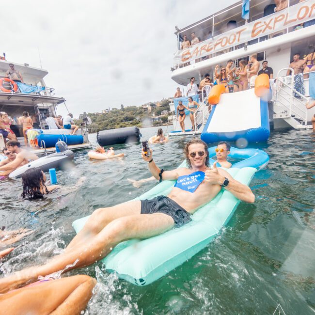 A man lounges on a blue pool float in the water, holding a drink and raising his other arm cheerfully. Several people swim around him, enjoying themselves near a large boat with a blue slide. The backdrop includes the shoreline and other boats, perfectly embodying The Yacht Social Club Event Boat Charters experience.