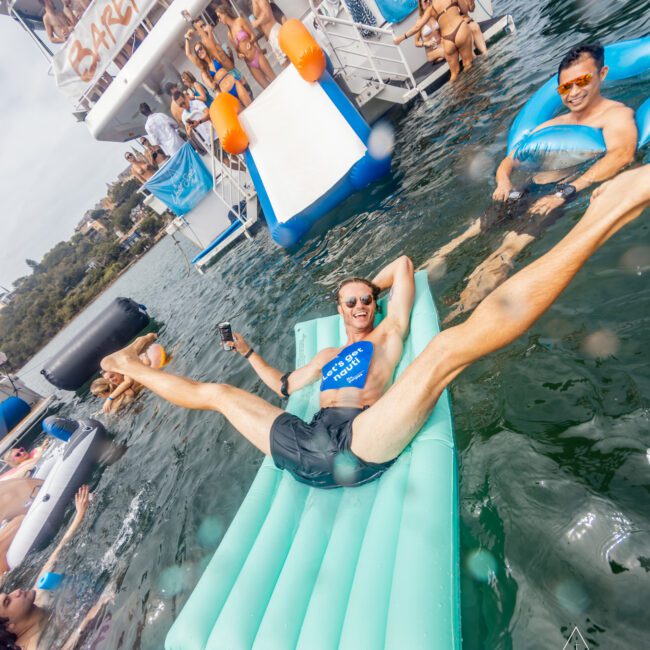 A man lounges on a mint green inflatable raft, holding a drink and wearing sunglasses, surrounded by others swimming and floating nearby. Behind him, people are on a boat with a slide leading into the water; a "Barefoot Boat" banner is visible. Perfect for Sydney Harbour Boat Hire The Yacht Social Club events.