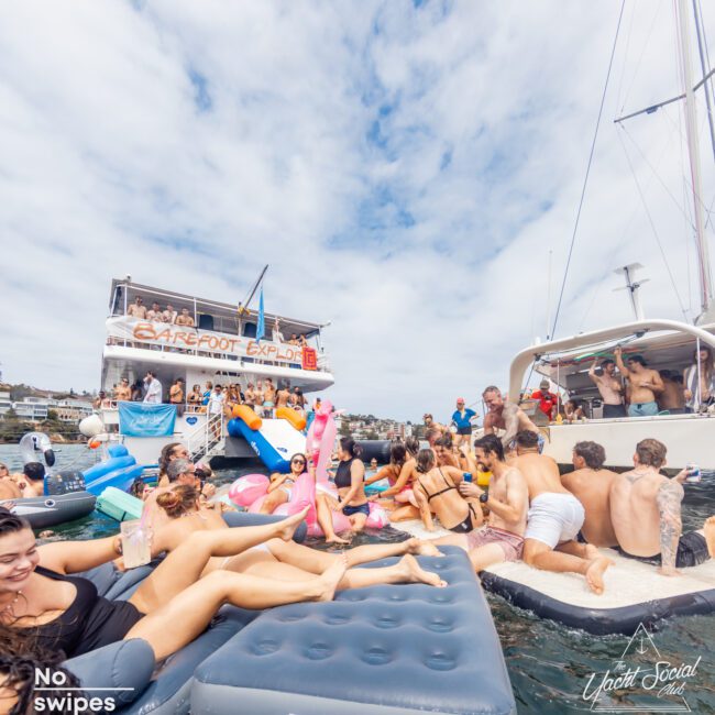 A lively scene of people enjoying a sunny day on the water. Several individuals are floating on inflatable loungers and air mattresses between two boats. The background features a large boat with "Barefoot Explorer" written on it, part of The Yacht Social Club Event Boat Charters. The sky is partly cloudy.