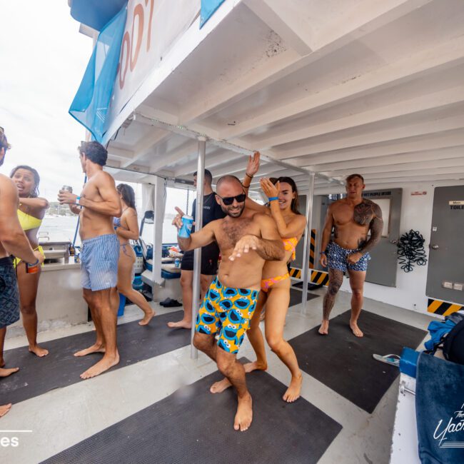 A lively group of people in swimsuits is dancing energetically on the deck of a boat. The setting is festive and cheerful, with men and women enjoying the sunny weather. The boat, part of "The Yacht Social Club Sydney Boat Hire," adds to the fun atmosphere with a visible banner displaying its name.