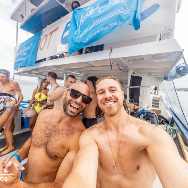 Two shirtless men smiling and taking a selfie on a boat with others in the background enjoying The Yacht Social Club Event Boat Charters. The boat has a banner reading "Yacht Social Club" and people in swimwear are visible. The scene is lively with a clear sky and water surrounding them.