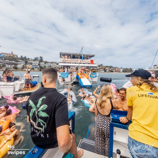 A vibrant scene of people enjoying a boat party on the water. Several boats from Sydney Harbour Boat Hire The Yacht Social Club are docked, and people are swimming, lounging on inflatable rafts, or interacting on the boats. An inflatable slide connects one boat to the water, adding to the festive atmosphere.