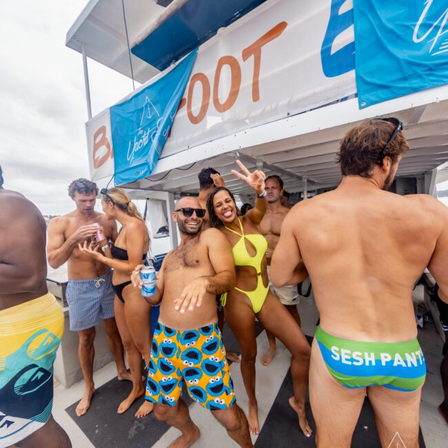 A group of people are partying on a boat in Sydney Harbour. They are wearing swimsuits and holding drinks, dancing and posing for the camera. A man in blue patterned shorts and a woman in a bright yellow swimsuit add to the lively, cheerful atmosphere at The Yacht Social Club Event Boat Charters.