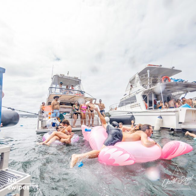 People are having fun in the water next to a couple of boats. Some are on inflatable floats, while others swim or relax on the boats. The lively and festive scene is part of The Yacht Social Club Sydney Boat Hire, with a cloudy sky overhead indicating a social and outgoing atmosphere.