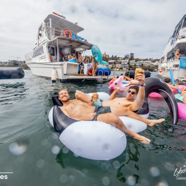 Two men relax on inflatable pool floats in the water, holding drinks and smiling towards the camera. Behind them, a group of people are on boats from The Yacht Social Club Sydney Boat Hire and an inflatable slide can be seen. The scene depicts a lively, social atmosphere on a sunny day by the water.