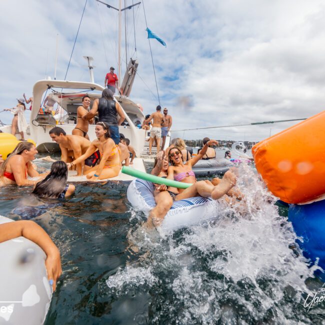 A lively group of people swim and float on inflatables near a boat. Splashing and playing, they enjoy the sunny day on the water, with some climbing back onto The Yacht Social Club Sydney Boat Hire. The sky is partly cloudy, adding a vibrant backdrop to the cheerful scene.