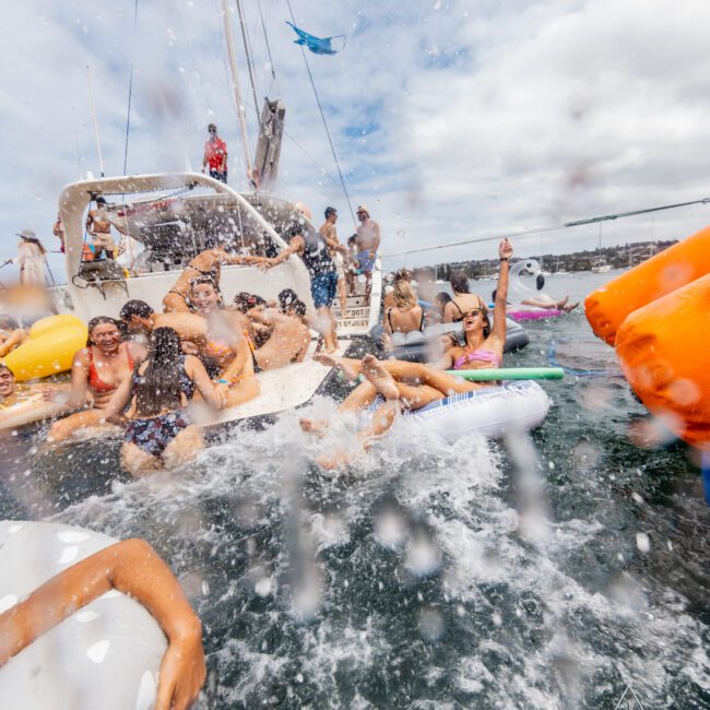 A group of people in swimsuits enjoying a lively party on a yacht. Some are sitting on the boat while others are in the water on inflatable floats. Water splashes in the foreground, and the sky is partly cloudy. Various float toys are visible at The Yacht Social Club Sydney Boat Hire.