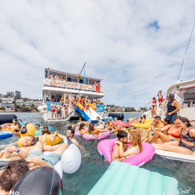 A lively party scene on water shows people enjoying themselves on colorful inflatable floats around a boat named "Barefoot Explorer." The luxury yacht rentals Sydney boat has a slide, and people are on the boat's deck and in the water, socializing under a partly cloudy sky.