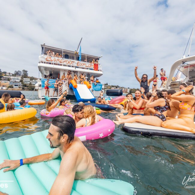 A lively scene of people enjoying a day on the water. Some are floating on inflatable devices, while others are gathered on a boat with slides and climbing structures. The sky is partly cloudy, and everyone is dressed in swimwear, laughing and having fun at The Yacht Social Club Event by Luxury Yacht Rentals Sydney.