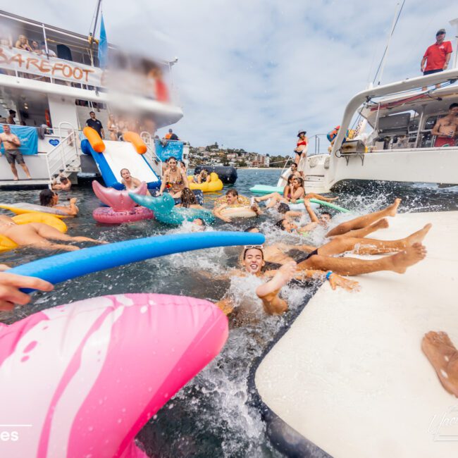 A lively scene of people swimming and floating on colorful inflatables around a boat named "Barefoot". Some people are playing with pool noodles, while others relax on the inflatables. The water splashes as swimmers move about under the clear sky. Experience unforgettable moments with Boat Parties Sydney The Yacht Social Club.