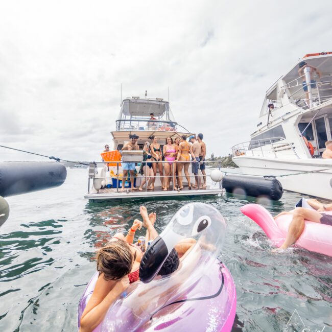 A group of people are gathered on the deck of a yacht, with two individuals in the foreground floating on inflatable pool toys in the water. The scene is lively with a cloudy sky above. This image represents The Yacht Social Club Sydney Boat Hire and features logos for "No Swipes" and "Yacht Social" in the corners.