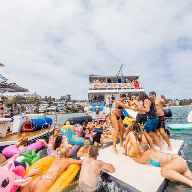 A lively group of people in swimwear are partying on a floating dock in the water, surrounded by inflatable toys. Boats and a party barge named "Barefoot & Fluid" are in the background. Many are dancing, lounging, and enjoying the sunny day at The Yacht Social Club Event Boat Charters.