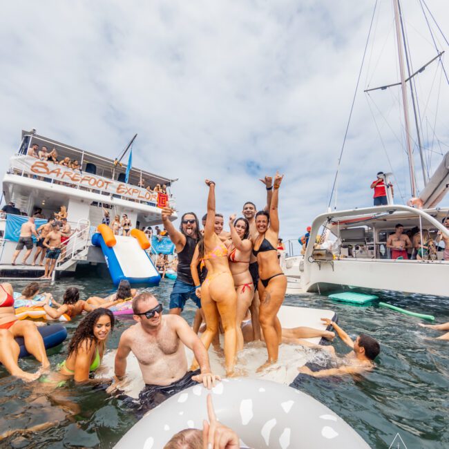 A group of people are posing and cheering on a large inflatable platform in the water. Several boats from The Yacht Social Club Sydney Boat Hire are nearby, with more people swimming and relaxing on floats. The sky is partly cloudy, creating a lively, fun atmosphere at a pool or beach party.