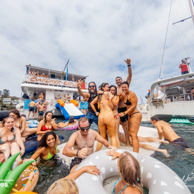 A lively group of people enjoys a sunny day on the water, surrounded by inflatables near a boat named "Barefoot Express," chartered through The Yacht Social Club Event Boat Charters. They cheer and pose for photos, with a mix of laughter and fun. The sky is partly cloudy, and other boats are visible in the background.