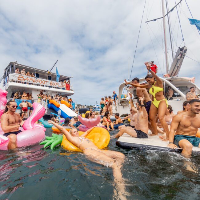 A lively group of people from The Yacht Social Club Sydney Boat Hire are enjoying a party on a boat and in the water. Some of them are on colorful floaties shaped like a flamingo, pineapple, and ice cream cone. Others are dancing or lounging on the boat. The sky is partly cloudy, and the atmosphere is festive.