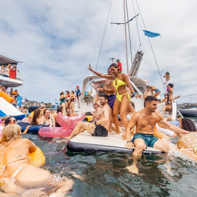 A lively party scene on a yacht docked in the water. People are dancing, swimming, and lounging on floaties. A woman in a yellow bikini poses on the edge of the yacht. The sky is partly cloudy and "The Yacht Social Club Sydney Boat Hire" is visible on the bottom right corner.