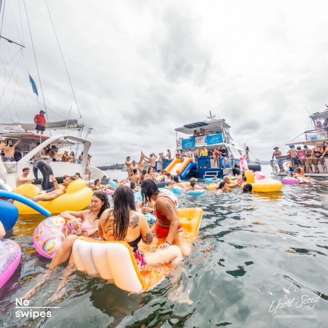 A vibrant scene of a boat party with numerous people in swimsuits enjoying themselves on large inflatable pool floats in the water. Boats are anchored nearby, and the sky is cloudy. People are socializing, swimming, and having fun. Text reads "The Yacht Social Club Event Boat Charters" and "Yacht Social.