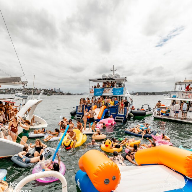 A lively boat party by The Yacht Social Club Sydney features numerous people enjoying themselves on inflatable floats and swimming near anchored yachts under a partially cloudy sky. The foreground has colorful inflatables, including boats and loungers, as people socialize and have fun.