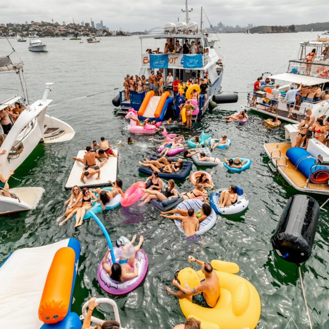 A vibrant scene of a party on the water with people enjoying themselves on inflatable rafts and pool toys. Several boats are anchored nearby, and one larger boat from The Yacht Social Club is in the center with people on board. The background features a city skyline under a cloudy sky.