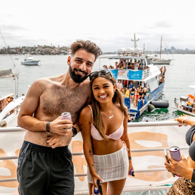 Two people are standing on a boat, smiling at the camera with a large boat full of people in the background. The man is shirtless, wearing black shorts and holding a drink. The woman is in a bikini top and skirt, holding water. Experience unforgettable moments with Sydney Harbour Boat Hire - The Yacht Social Club!