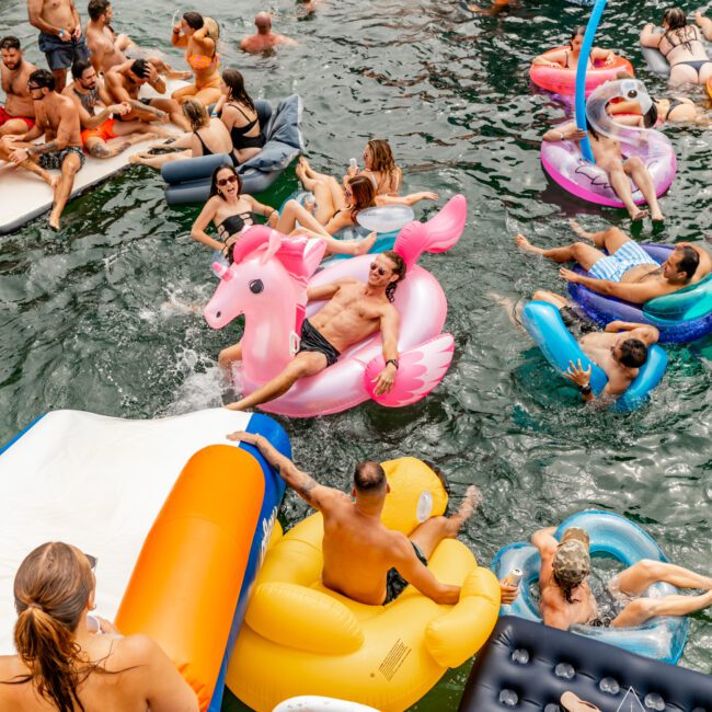 A lively scene of adults relaxing and having fun on various colorful inflatables in the water. People are sunbathing, chatting, and enjoying the summer day by The Yacht Social Club Sydney Boat Hire. The water is crowded with an array of floaties, including unicorns, ducks, and loungers.