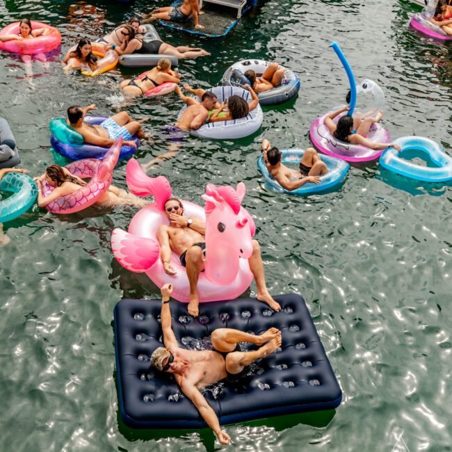 A group of people enjoy a day on the water, lounging on inflatable floats like a flamingo, unicorn, and donut. They swim and relax near a docked boat in sunny weather. For an even grander experience, check out Luxury Yacht Rentals Sydney or boat parties with The Yacht Social Club.