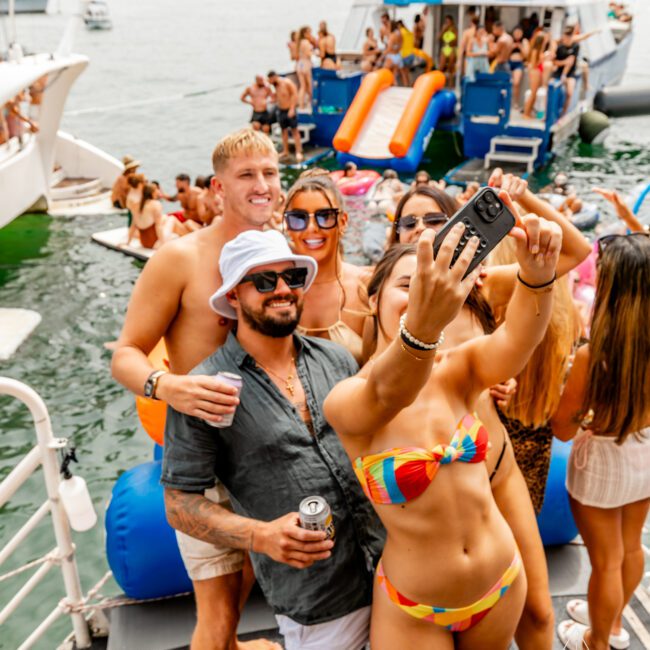 A group of people wearing swimsuits smile and pose for a selfie on a sunny day aboard a boat covered with inflatables and pool toys. Other yachts and people in the water are visible in the background, showcasing the vibrant scene at Boat Parties Sydney The Yacht Social Club. Everyone seems to be having a fun, lively time.