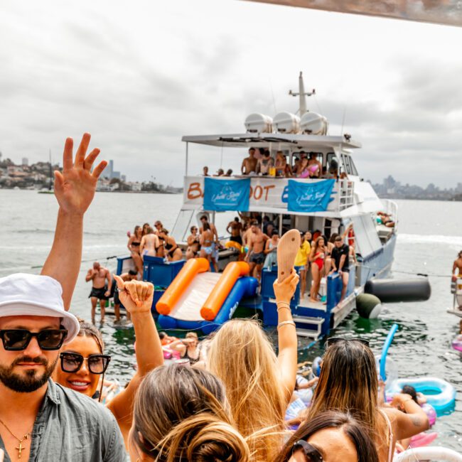 A lively boat party on a sunny day, with people in swimwear enjoying music, dancing, and inflatable slides. The scene includes a crowded yacht from Luxury Yacht Rentals Sydney in the background, a man in a white hat, and another wearing sunglasses. The water is calm and inviting.