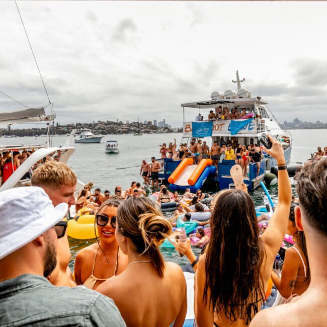 A lively boat party on the water featuring numerous people in swimwear. Partygoers are mingling on multiple boats with inflatable floats, slides, and rafts. The scene is vibrant with socializing and activities, set against a cloudy sky and distant skyline at The Yacht Social Club Sydney Boat Hire.