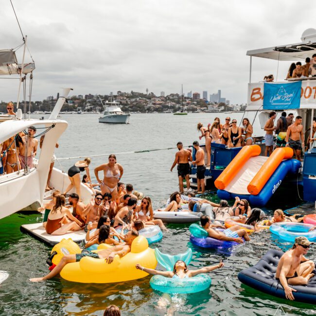 A lively boat party hosted by The Yacht Social Club is taking place on a sunny day in Sydney. People are swimming and relaxing on colorful inflatables, while others enjoy a slide from the boat into the water. The scene includes several boats and a cityscape in the background, perfect for Boat Rental and Parties.