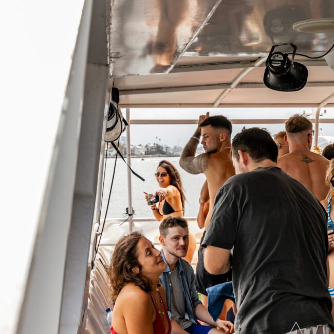 A group of people are socializing on a boat from Sydney Harbour Boat Hire The Yacht Social Club. Some are standing and some are seated, with a woman in a red dress talking to a man in a gray t-shirt. In the background, a shirtless man with tattoos and a woman in a black bikini are visible.