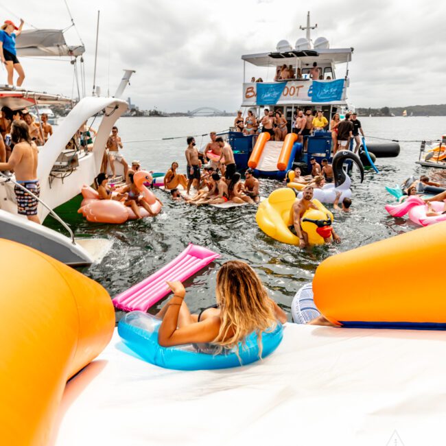 A lively scene of people at a boat party in the water. Many are on colorful floaties such as flamingos and lounge chairs. The sky is overcast, and everyone enjoys the festive atmosphere. In the background is a large boat with more people onboard, showcasing The Yacht Social Club Sydney Boat Hire experience.