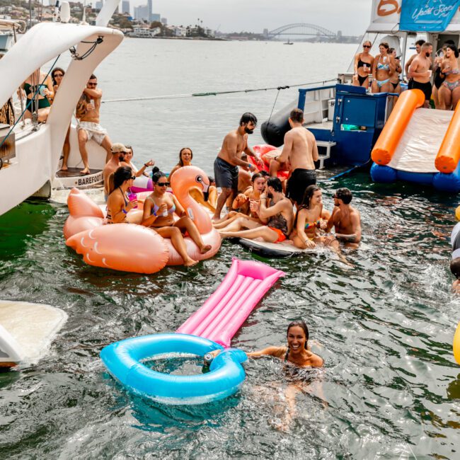 A lively scene of people enjoying a boat party hosted by The Yacht Social Club. Some are on the deck and some in the water, floating on inflatable pool toys like a pink flamingo and blue ring. A woman in the water smiles with her arms on a pink float. The shoreline of Sydney Harbour is visible in the background.