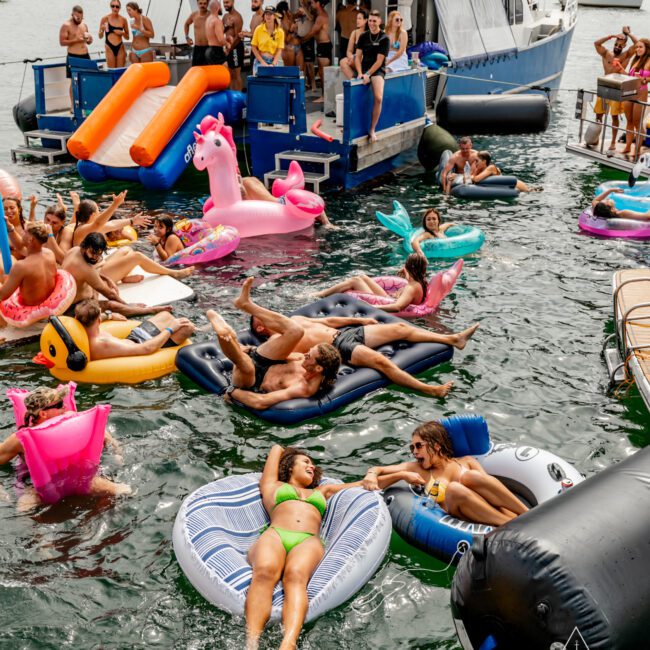 A lively group of people enjoying a sunny day on inflatable floats in the water beside a yacht, courtesy of Sydney Harbour Boat Hire The Yacht Social Club. They are lounging and socializing on colorful floats like flamingos, donuts, and lounge chairs. Some are on the yacht, while others are in the water.