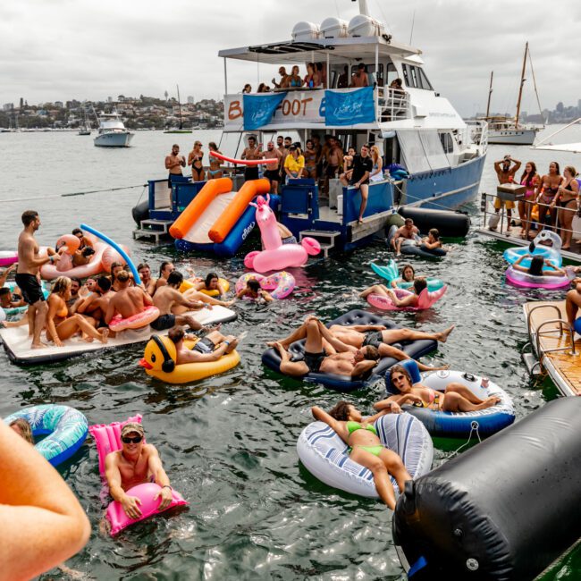 A lively scene of people enjoying a party on inflatable floats in the water, gathered around boats. Various inflatables, including flamingos, unicorns, and pool mattresses, are occupied by partygoers in swimsuits under a cloudy sky. Experience boat parties with The Yacht Social Club Sydney Boat Hire.