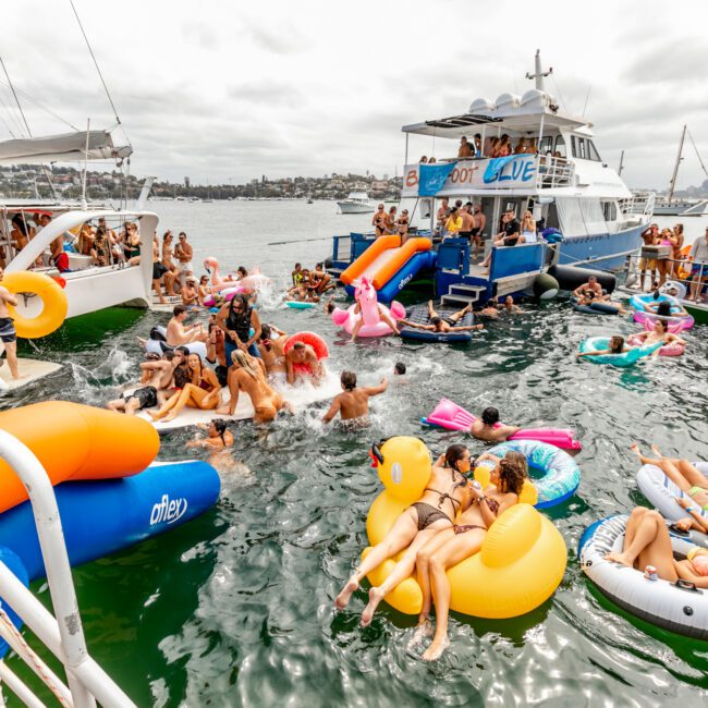 A lively scene of people enjoying a party on boats and in the water, with colorful inflatable pool floats. Several boats from The Yacht Social Club Sydney Boat Hire are docked together, and people are swimming and socializing between them. The background shows a cloudy sky and distant land.