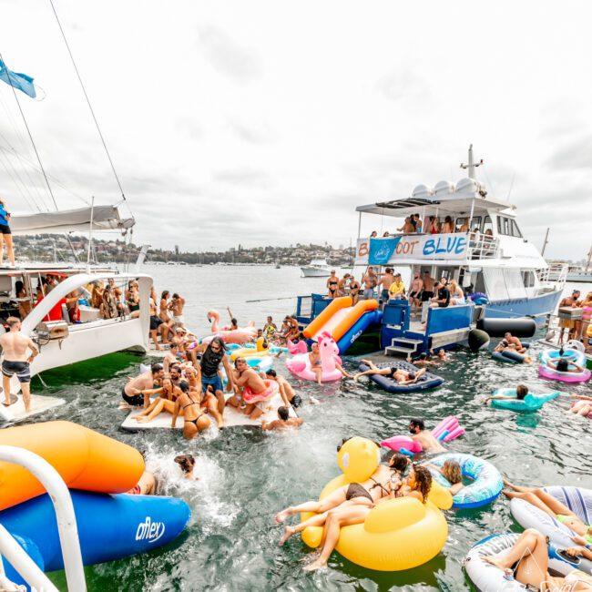 A lively scene at a boat party hosted by The Yacht Social Club, with people on and around multiple boats, some sliding down colorful water slides into the water. Others are floating on inflatables and swimming. The event appears festive with clear skies and a city skyline in the background.