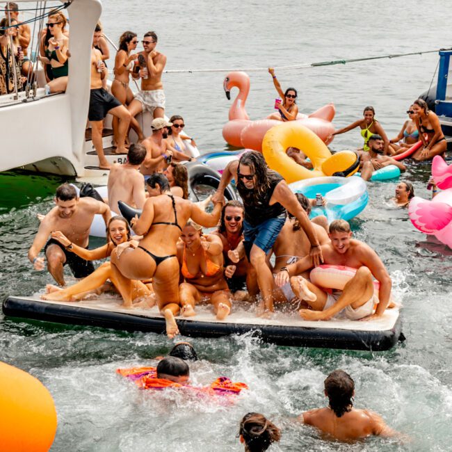 A lively scene on a sunny day with numerous people on a docked boat and in the water. Many are on inflatables, including a unicorn and flamingo float. A group is gathered on a large float, enjoying the water and sun. The background features a city skyline, highlighting The Yacht Social Club Sydney Boat Hire experience.